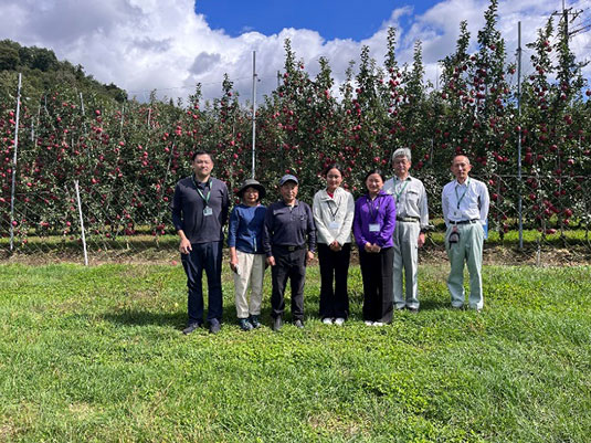 3．高密度栽培のリンゴ園を背に研修関係者の皆さんとの集合写真