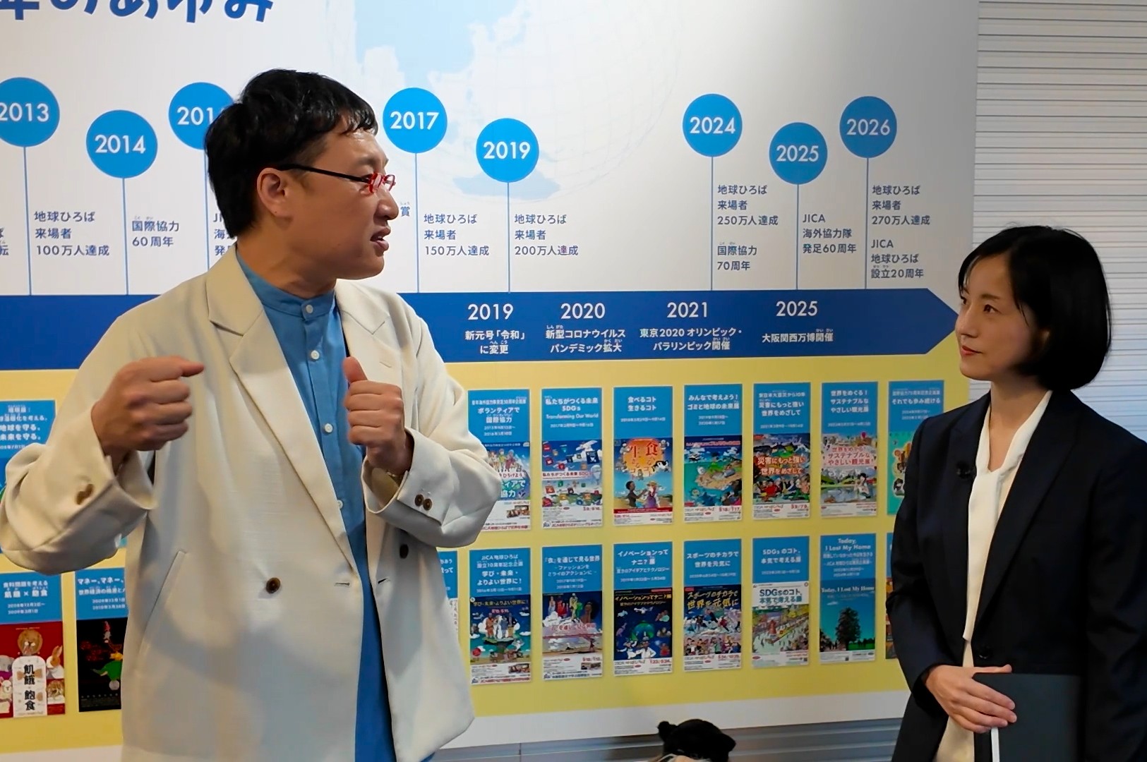 年表で20年を振り返り、山里亮太さんに地球ひろばの歩みを説明する植田茜・JICA広報部地球ひろば推進課課長（右）