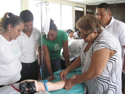 静脈注射：看護教員が学生に供与機材である静脈注射用シュミレーターを使用し、静脈注射の方法を説明している様子。