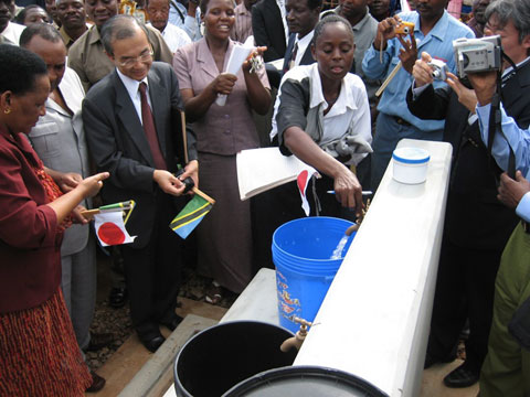 動力ポンプ井戸給水施設（公共水栓）。貯水槽に溜めた地下水は水道管により公共水栓まで配られる。蛇口をひねれば安全な水を汲むことができる（2007年3月27日の第3期供与式典に出席された伊藤大使およびタンザニア水省副大臣）。